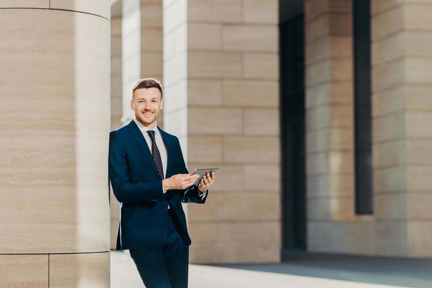Positive successful male entrepreneur in luxury black suit, holds modern tablet, looks happily somewhere, stands in modern office building, searches internet or social networks during work break photo