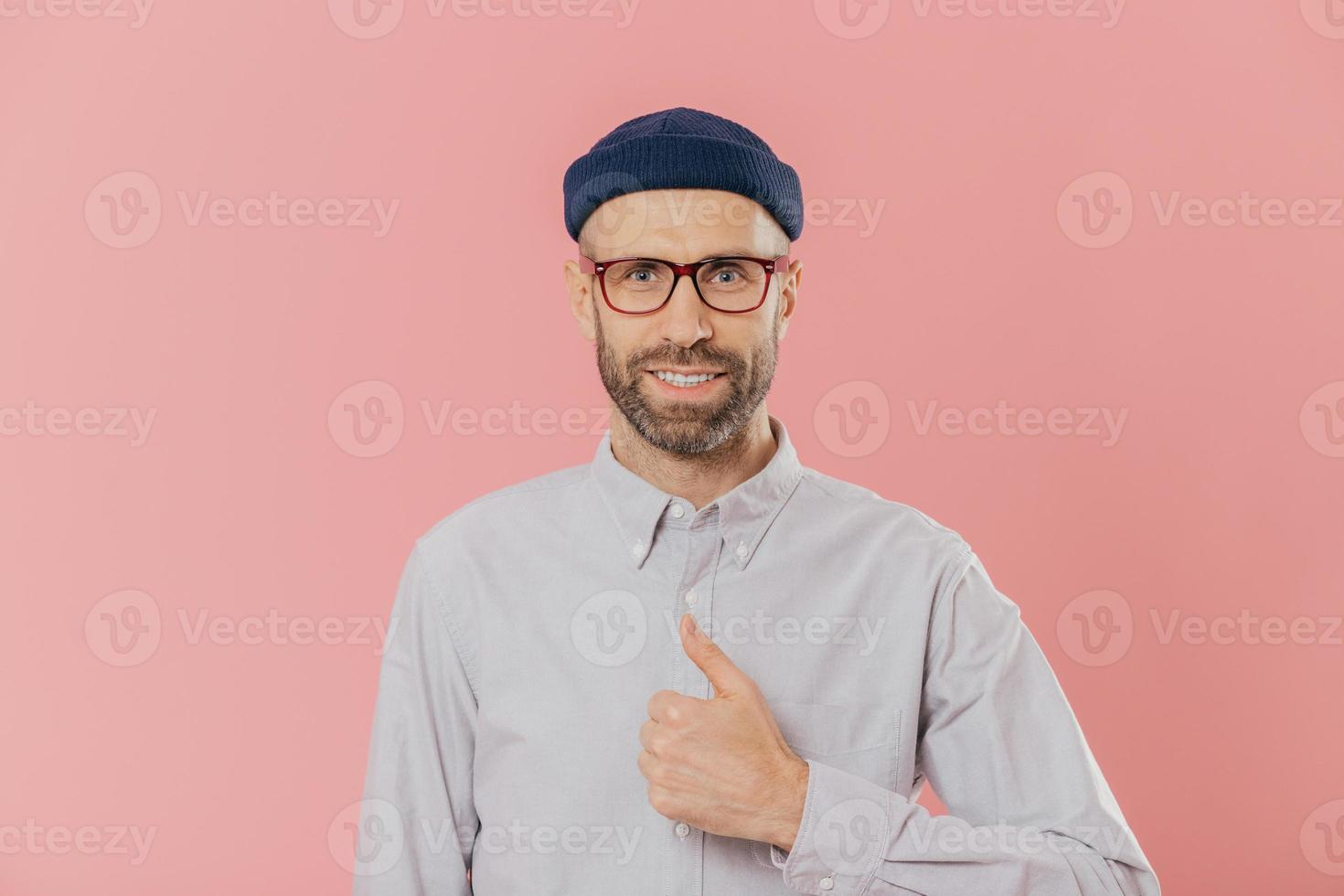 el hombre atractivo satisfecho tiene un aspecto atractivo, levanta el pulgar, está de acuerdo con algo, sonríe alegremente, usa sombrero y camisa, aislado sobre fondo rosa. guapo estudiante aprueba amigos buena idea foto