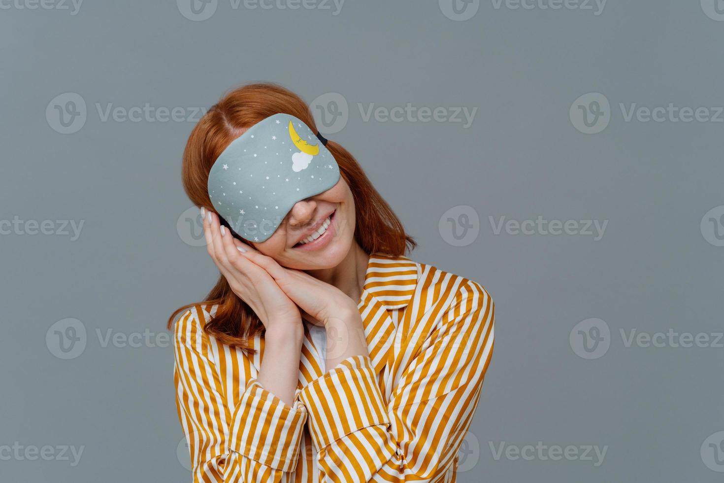 Just relaxing or time for nap. Smiling gentle woman with eye mask, tilts head on pressed palms, wears striped pajama, poses over grey wall, copy space area for your text feels comfortable and relaxed photo