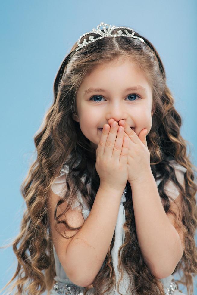 Image of positive beautiful smiling child keeps both hands on mouth, has long thick hair, blue eyes, wears crown, poses against blue background, expresses happiness. Children and satisfaction photo