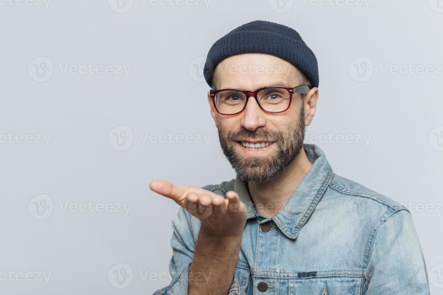 Glad bearded male in trendy eyewear expresses his love, shows air kiss, has cheerful expression, isolated over grey background. Blue eyed middle aged man in stylish clothing, gestures indoor photo