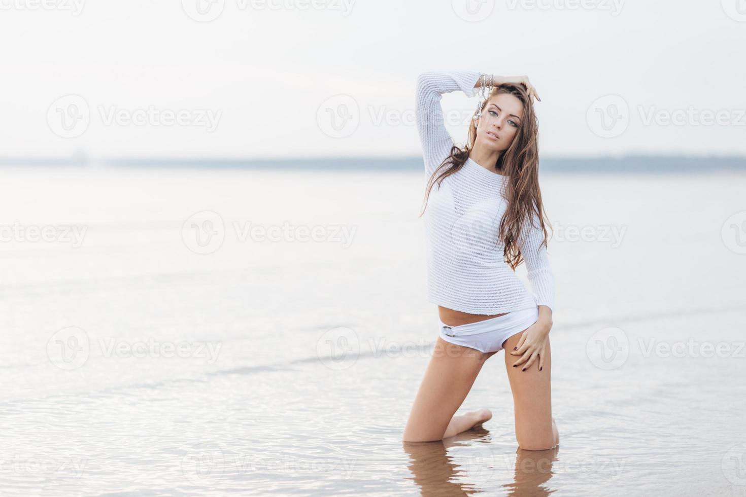 Attractive young female model models at coastline against beautiful sea view and clear sky, wears bikini, stands on knees, enjoys wonderful nature and loneliness. Perfect body shape and fitness photo