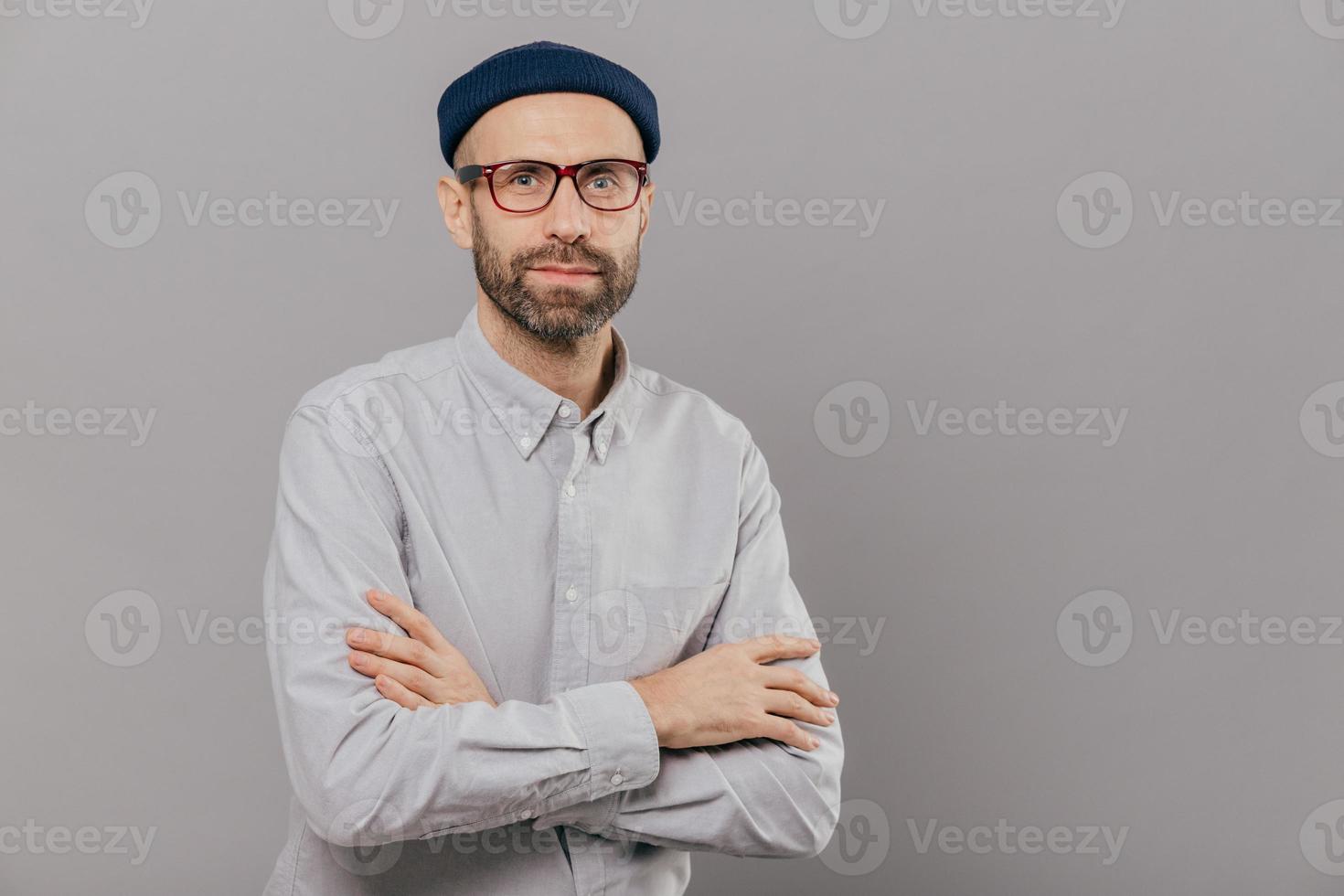 Photo of serious male keeps arms folded over chest, dressed in white formal shirt, fashionable hat, poses for making photography over grey wall with free space for your advertisement or promotion
