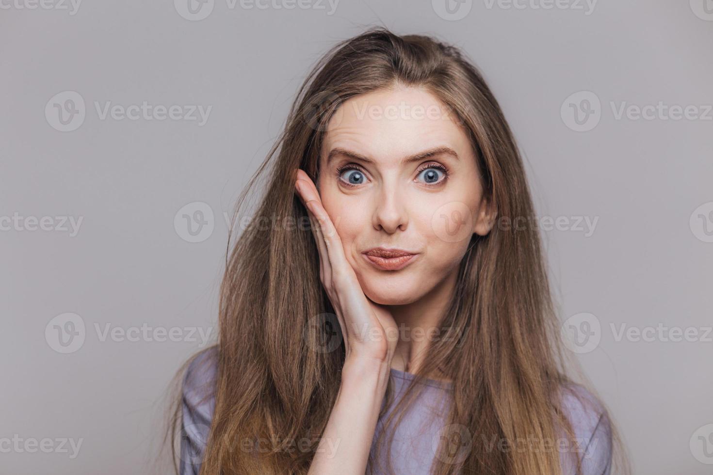 Attractive blue eyed brunette female looks with bewilderment at camera, has specific appearance, cant beleive in eyes, isolated over grey studio background. People and facial expressions concept photo