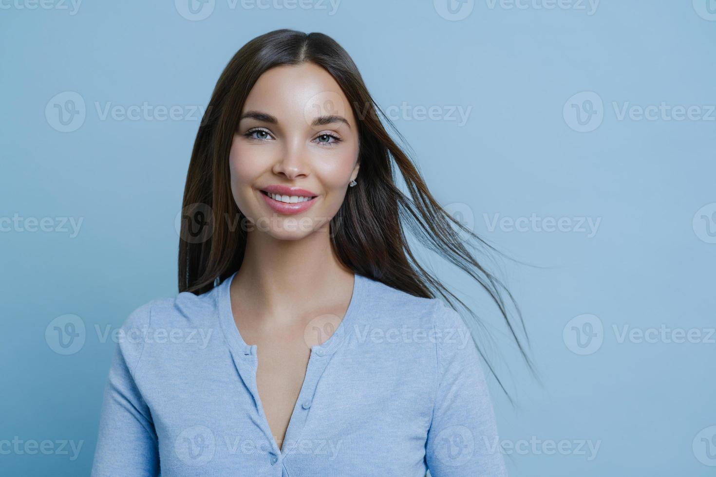 Positive lovely feminine lady happy to have first date with boyfriend, smiles broadly, has appealing appearance, glad to hear something pleasant, models indoor alone, wears blue casual jumper photo