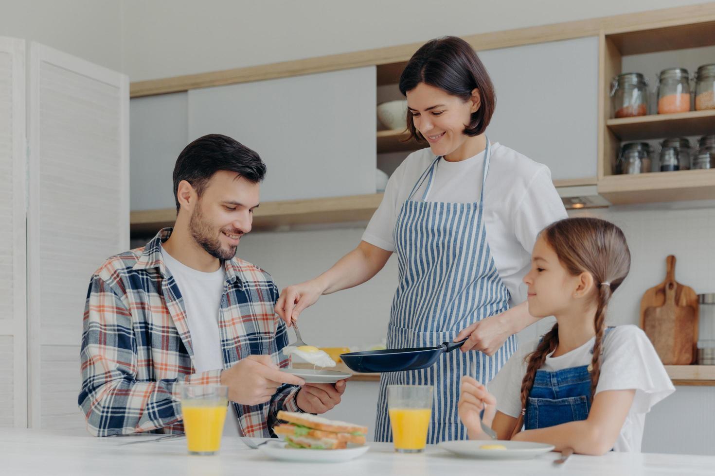 ama de casa ocupada sostiene una sartén, da comida preparada a marido e hija, desayuna juntos, se sienta en la mesa de la cocina con un vaso de jugo y un sándwich. familia feliz comer en casa foto