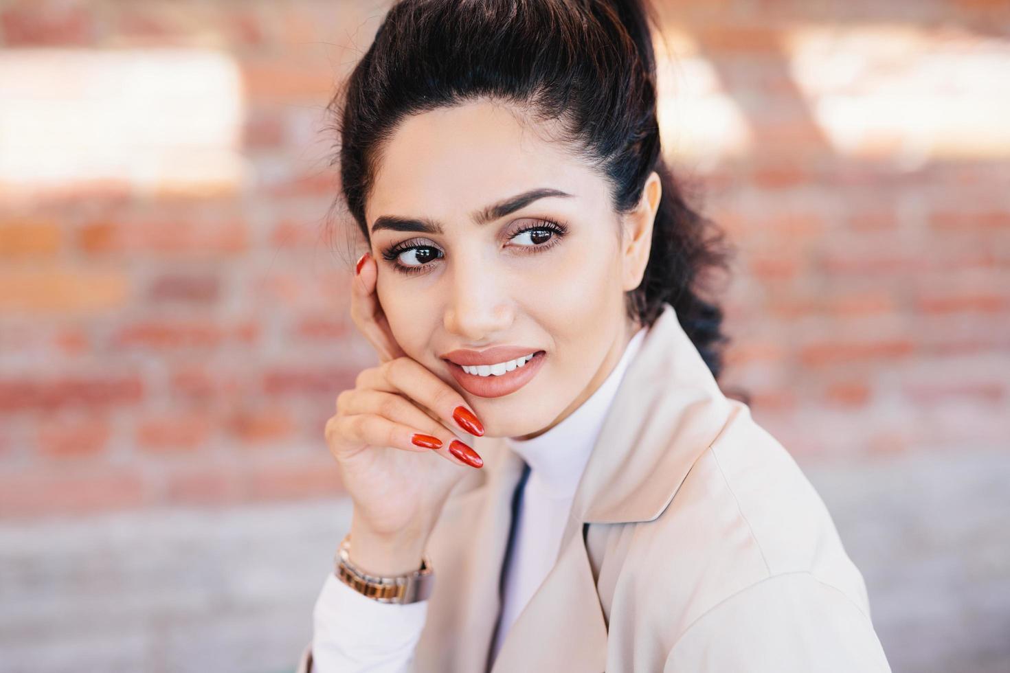 Close up of glamour brunette female having thin eyebrows, dark eyes with long eyelashes, pure skin, full well-shaped lips, red manicure wearing elegant watch on hand and white jacket looking aside photo