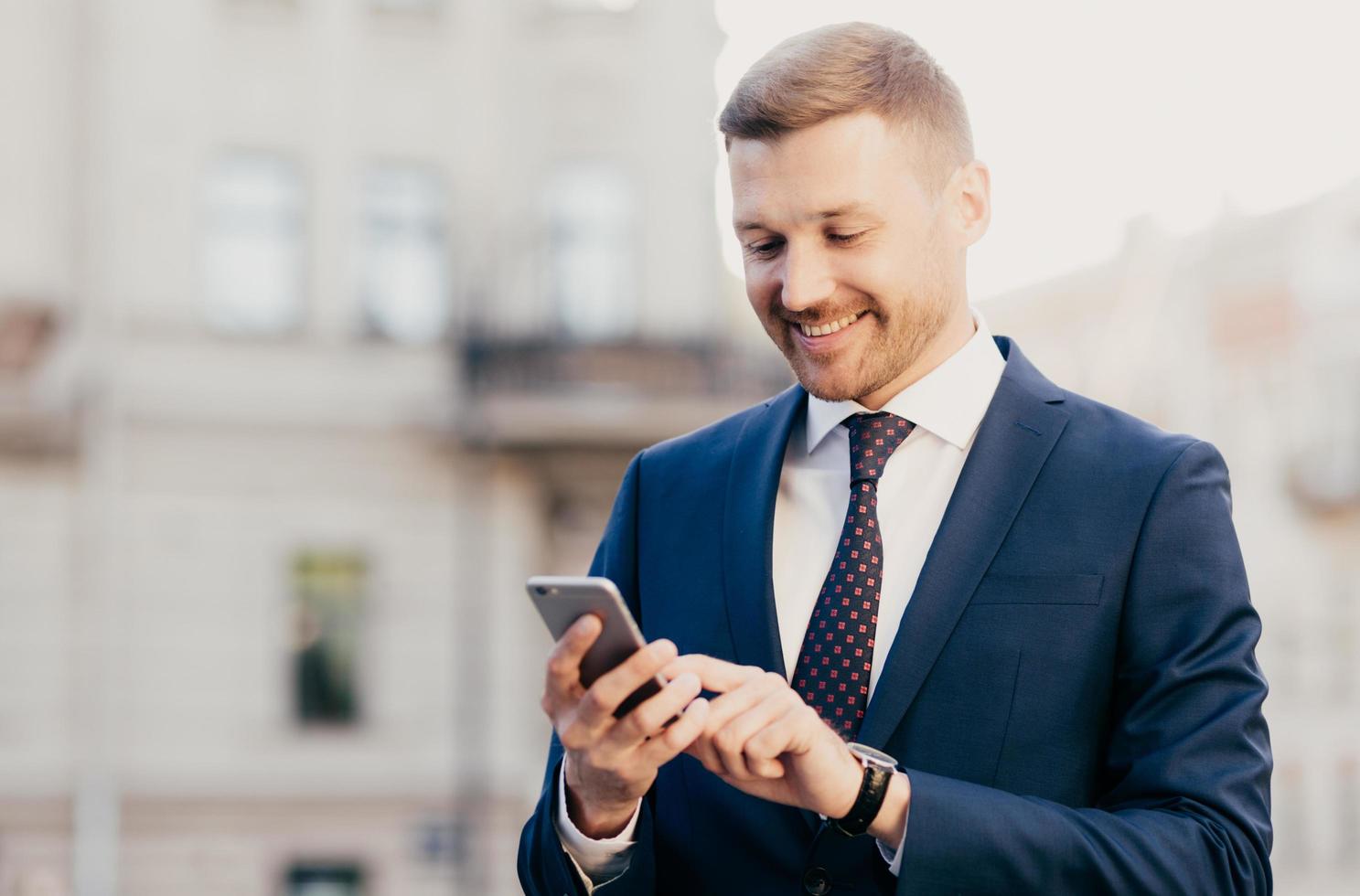 Intelligent businessman with positive expression, uses application on smart phone for navigating in big city, has working trip, wears elegant clothes, has happy look. People and occupation concept photo