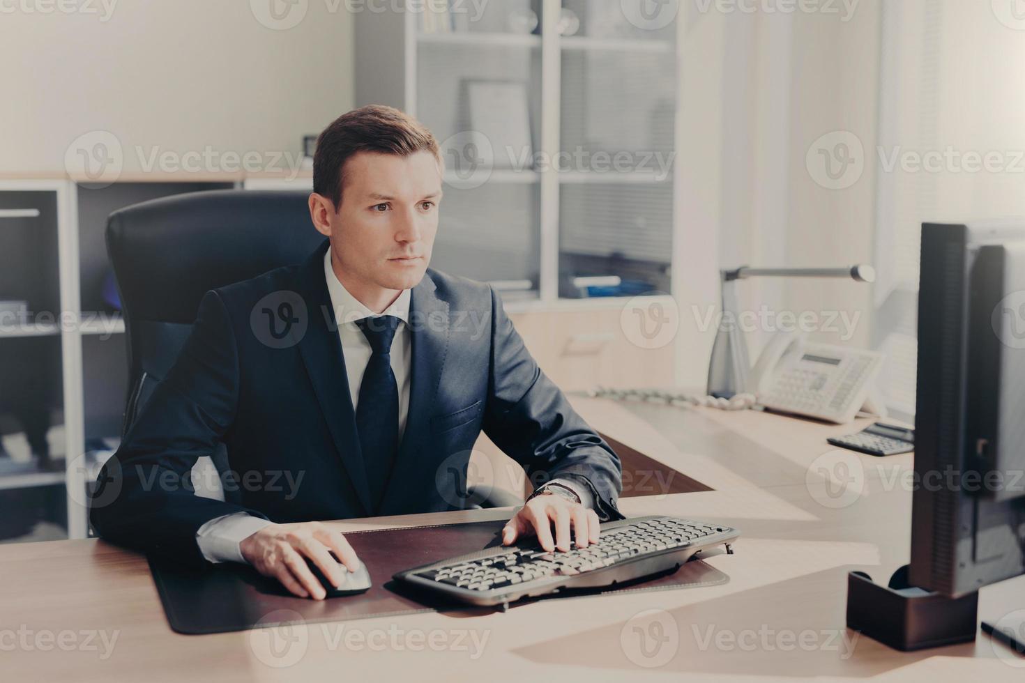 Serious concentrated businessman sits in front of computer at work place, keyboards information, wears suit, prepares business project for next year, poses in spacious cabinet. People and finances photo