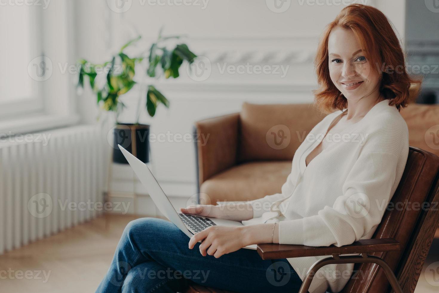Sideways shot of happy redhead young woman sits in comfortable armchair with laptop computer, works remotely, checks email and reads notification, browses useful website and installs application photo