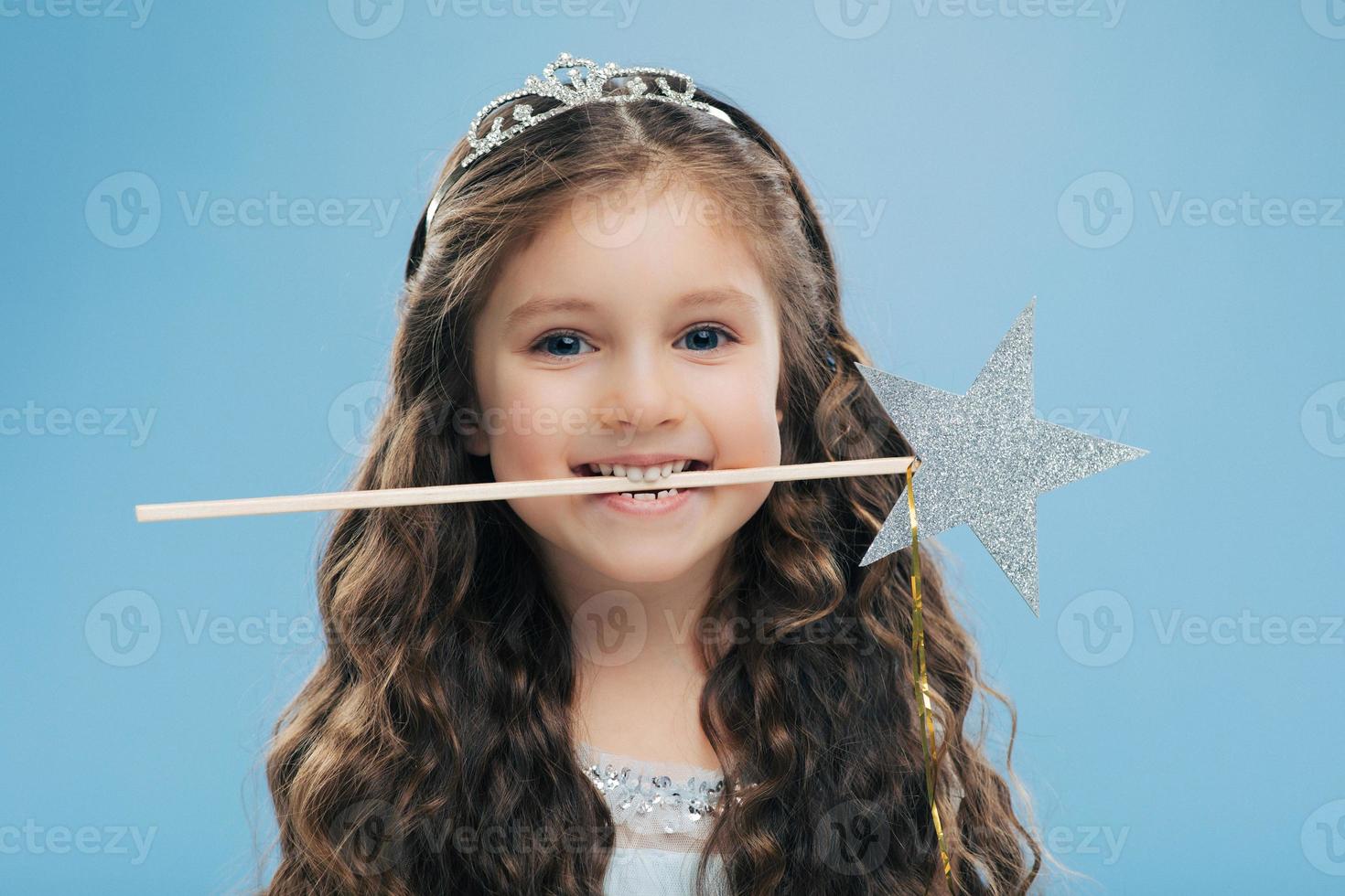 una foto horizontal de un niño pequeño y atractivo y sonriente sostiene una varita mágica en la boca, tiene el pelo castaño rizado, posa sobre un fondo azul, mira directamente a la cámara, lleva una corona. niños y concepto mágico