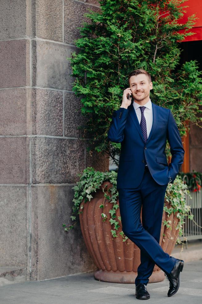 Clever handsome male manager in formal clothes, shares good news with business partner, being glad after successful meeting, wears formal clothes, stands near office building. Technology concept photo