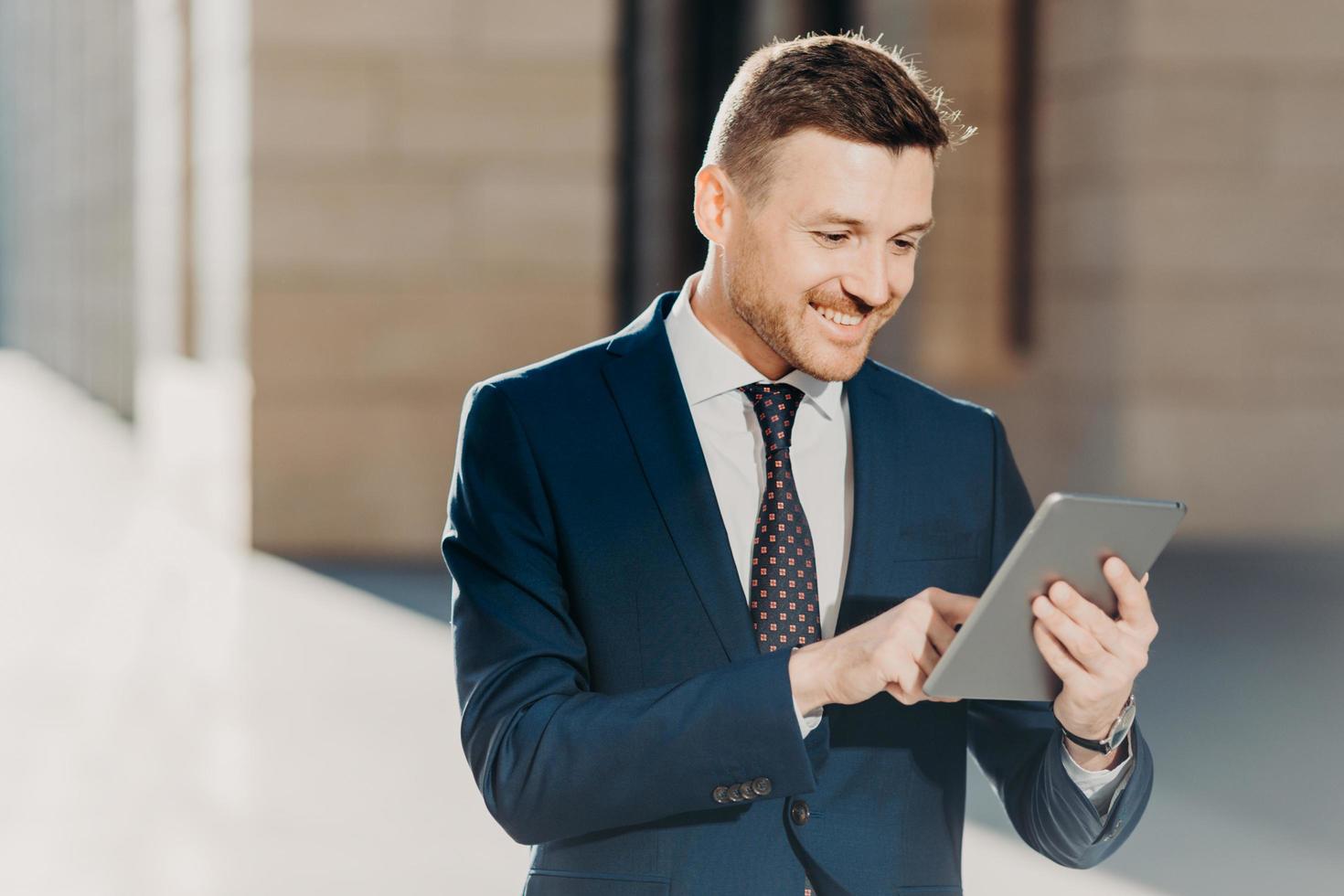 Succesful male entrepreneur in formal suit, holds modern tablet, checks documentation online, reads necessary information for making business report, uses free wifi connection. People and technology photo