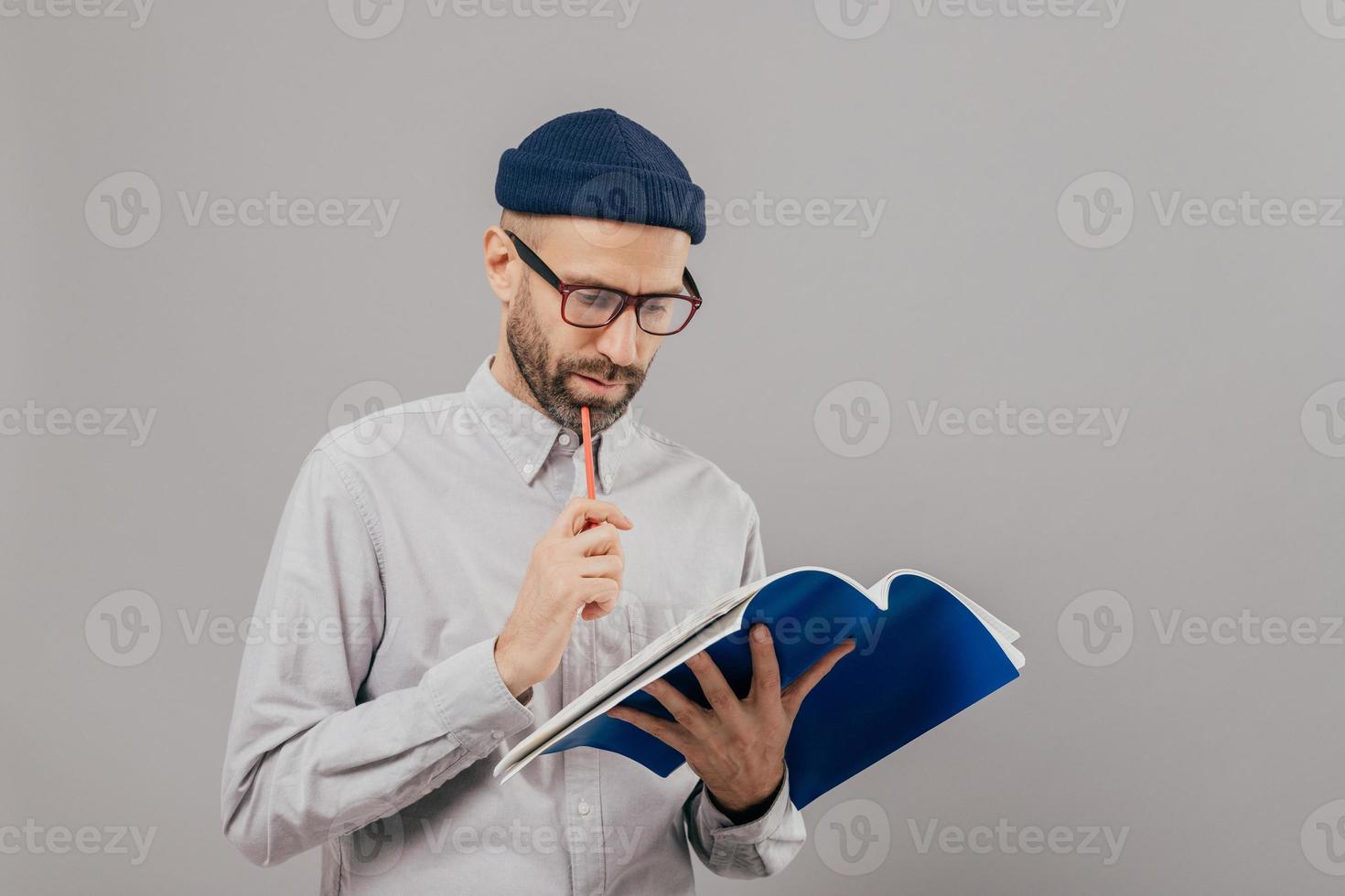 el hombre concentrado sin afeitar sostiene la pluma, mira atentamente en el libro abierto, aprende nueva información, se prepara para el seminario, usa ropa formal, aislado sobre fondo gris. tiempo para estudiar y aprender foto