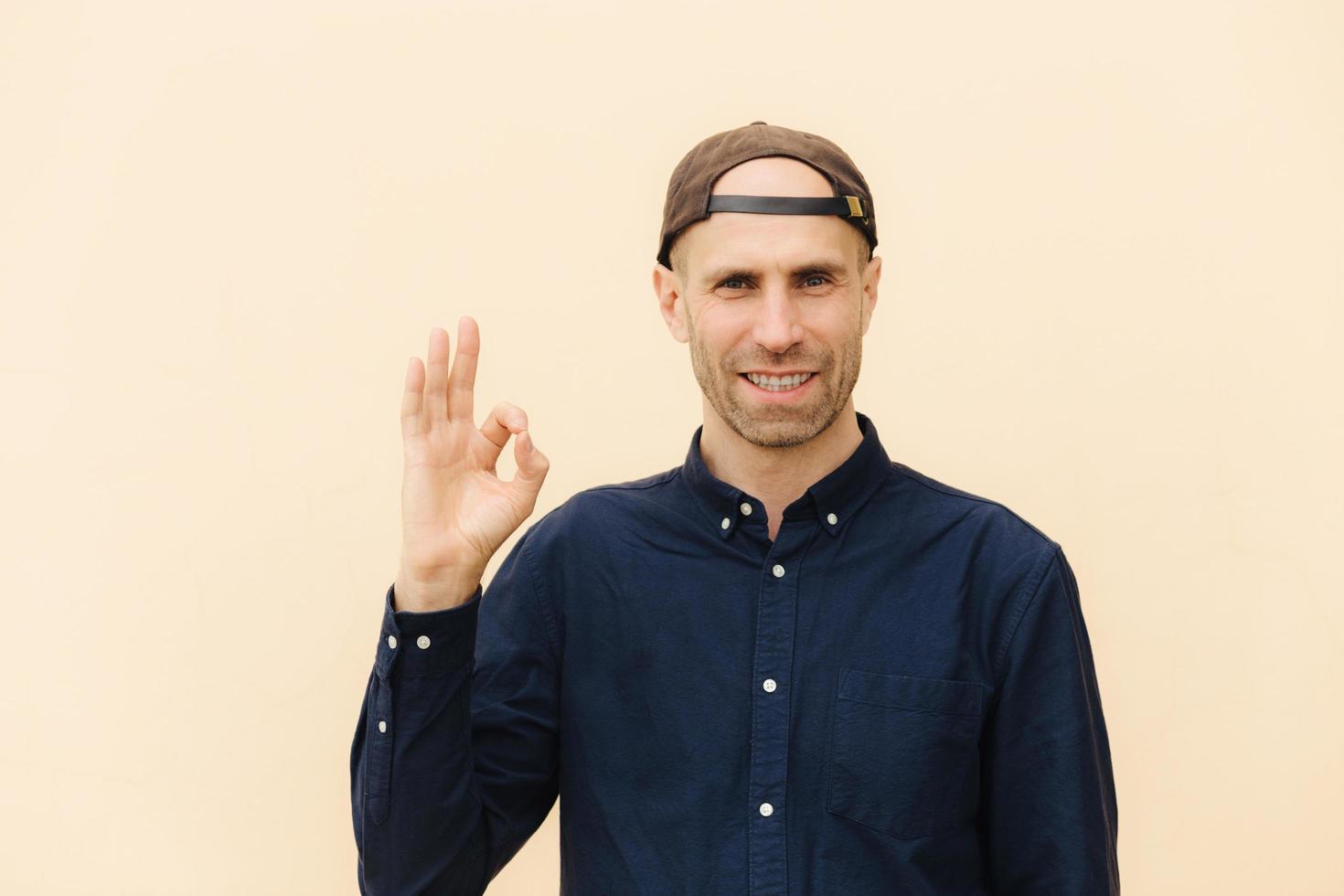 Everything is ok and fine Happy young man in shirt, gestures indoor, dressed in elegant shirt and fashionable cap, shows his approval, being satisfied with something, isolated over studio wall photo