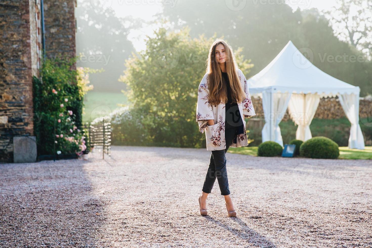Portrait of pleasant looking slim tall woman wears cape and high heeled shoes, stands crossed legs, poses outdoors against ancient area, has confident look directly into camera. Lifestyle concept photo