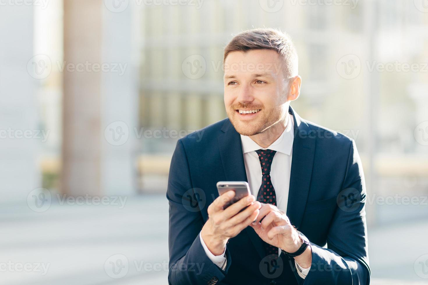 Delighted thoughtful elegant male marketing trader in formal black suit, uses modern cell phone for searching website in internet, types text message, looks pensively into distance. Bussiness concept photo