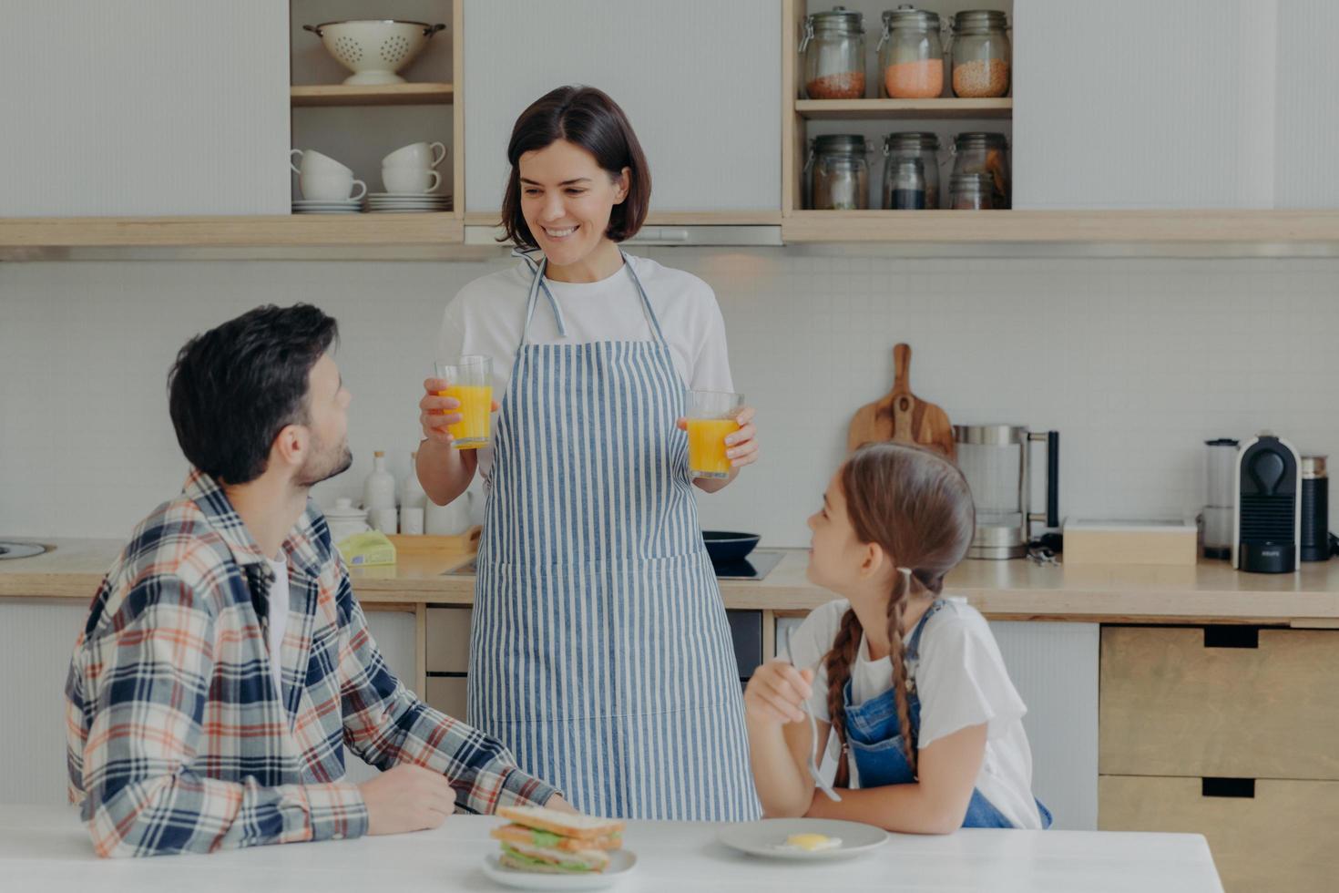 Lovely happy mother wears apron, holds two glasses of orange juice, talks to husband and daugher, prepares breakfast. Three membered family have dinner time at kitchen. Togetherness concept. photo
