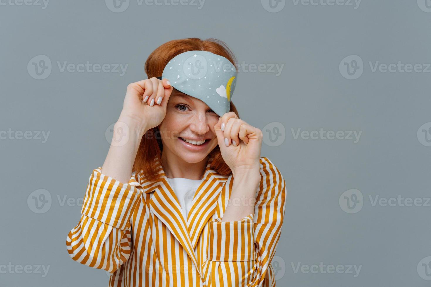 Playful woman with ginger hair looks from sleep mask, wears striped pajamas, has cheerful expression, poses over grey background, enjoys sleeping at home. Healthy sleep and relaxation concept photo
