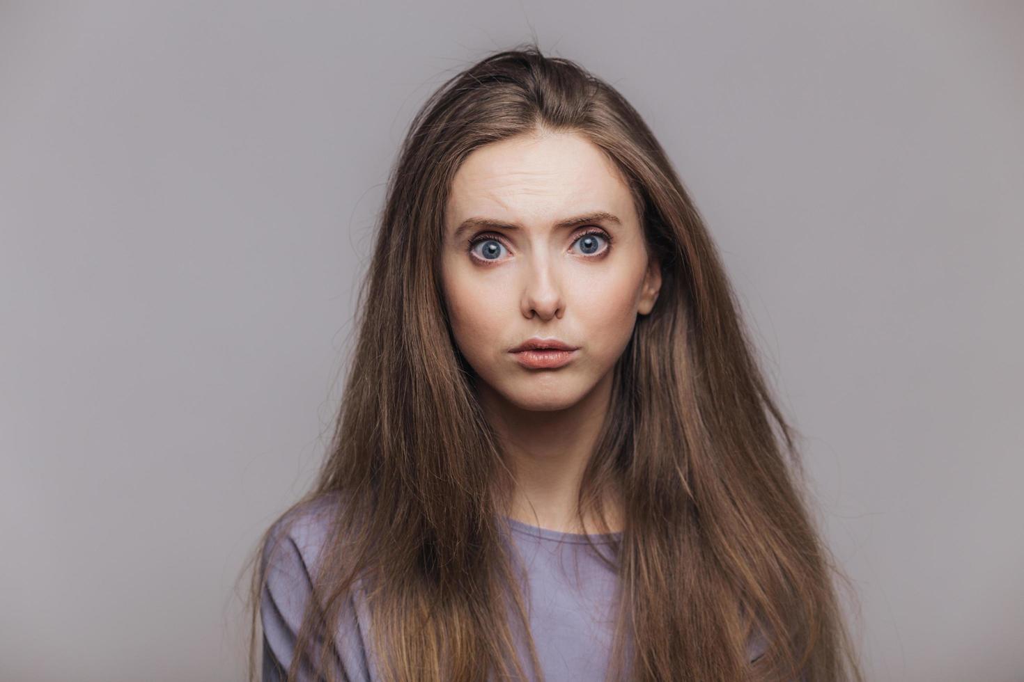 Studio shot of pleasant looking blue eyed female model with dark long hair, has puzzled expression as hears shocking news from interlocutor, isolated over grey background. Facial expressions concept photo