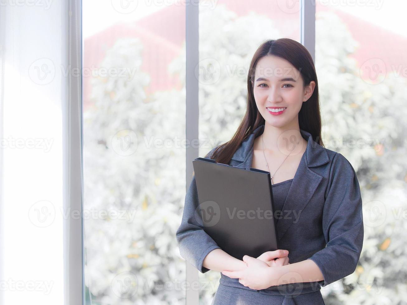 una joven empresaria asiática profesional viste un vestido gris y sonríe alegremente mientras trabaja y sostiene el portapapeles con confianza en el cargo. foto