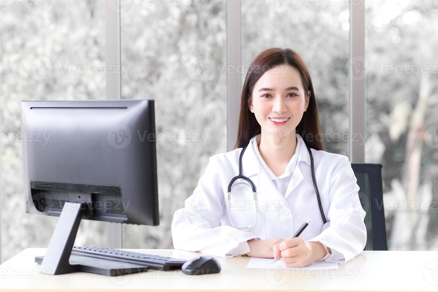 Asian beautiful woman doctor who wears medical coat is working in office room at hospital while has computer put on table. photo