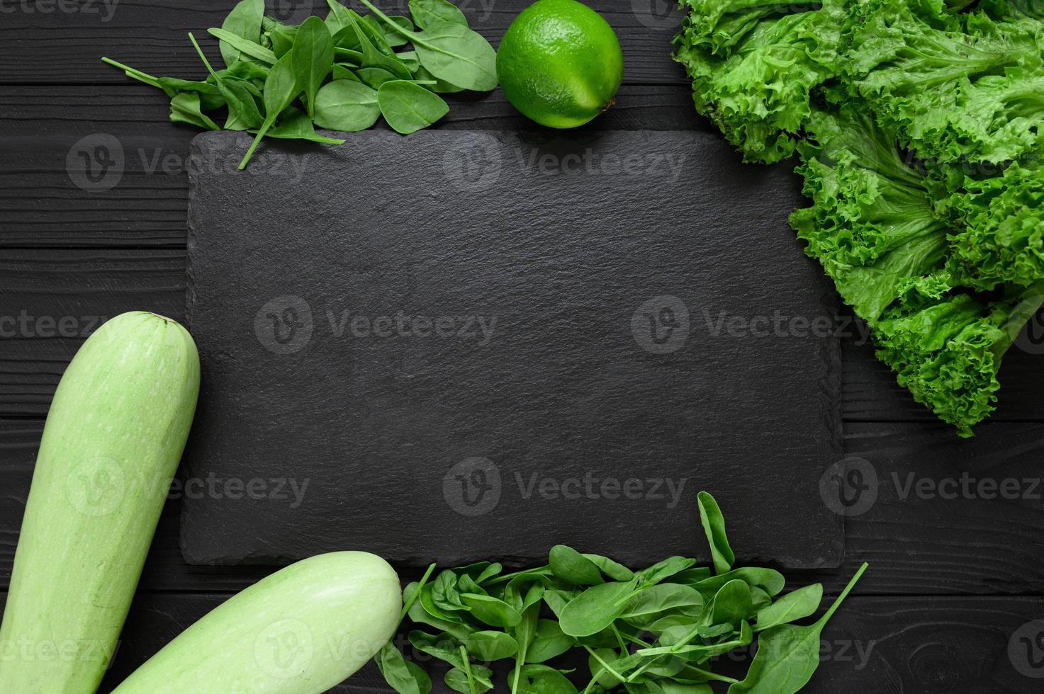 Black stone board on a black wooden background decorated with green vegetables and herbs. photo