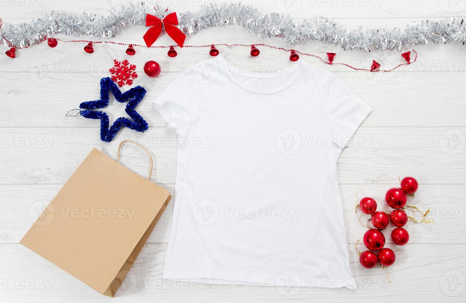 Close up white blank template t shirt with copy space paper bag. Christmas Holiday concept. Top view mockup t-shirt, star, white wooden background. Happy New Year decorations accessories. Xmas outfit photo