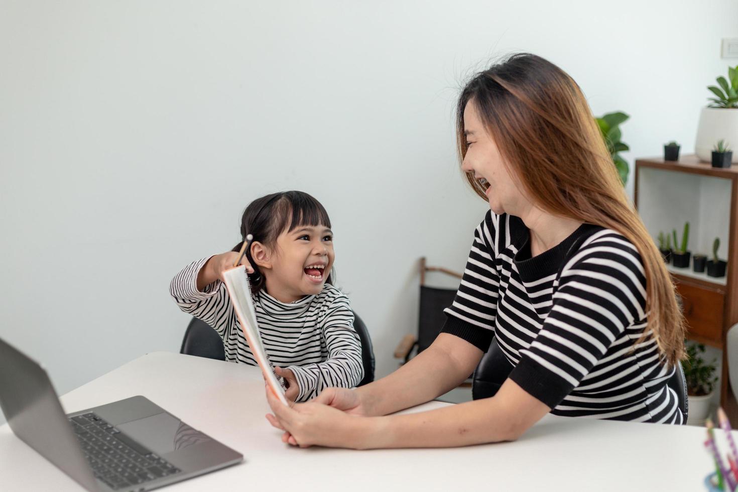 Niña pequeña asiática aprendiendo clases en línea en casa con su madre. el niño en edad preescolar usa una computadora portátil para hacer la tarea, la escuela en casa del maestro de la escuela por internet remoto digital con el apoyo de mamá. foto