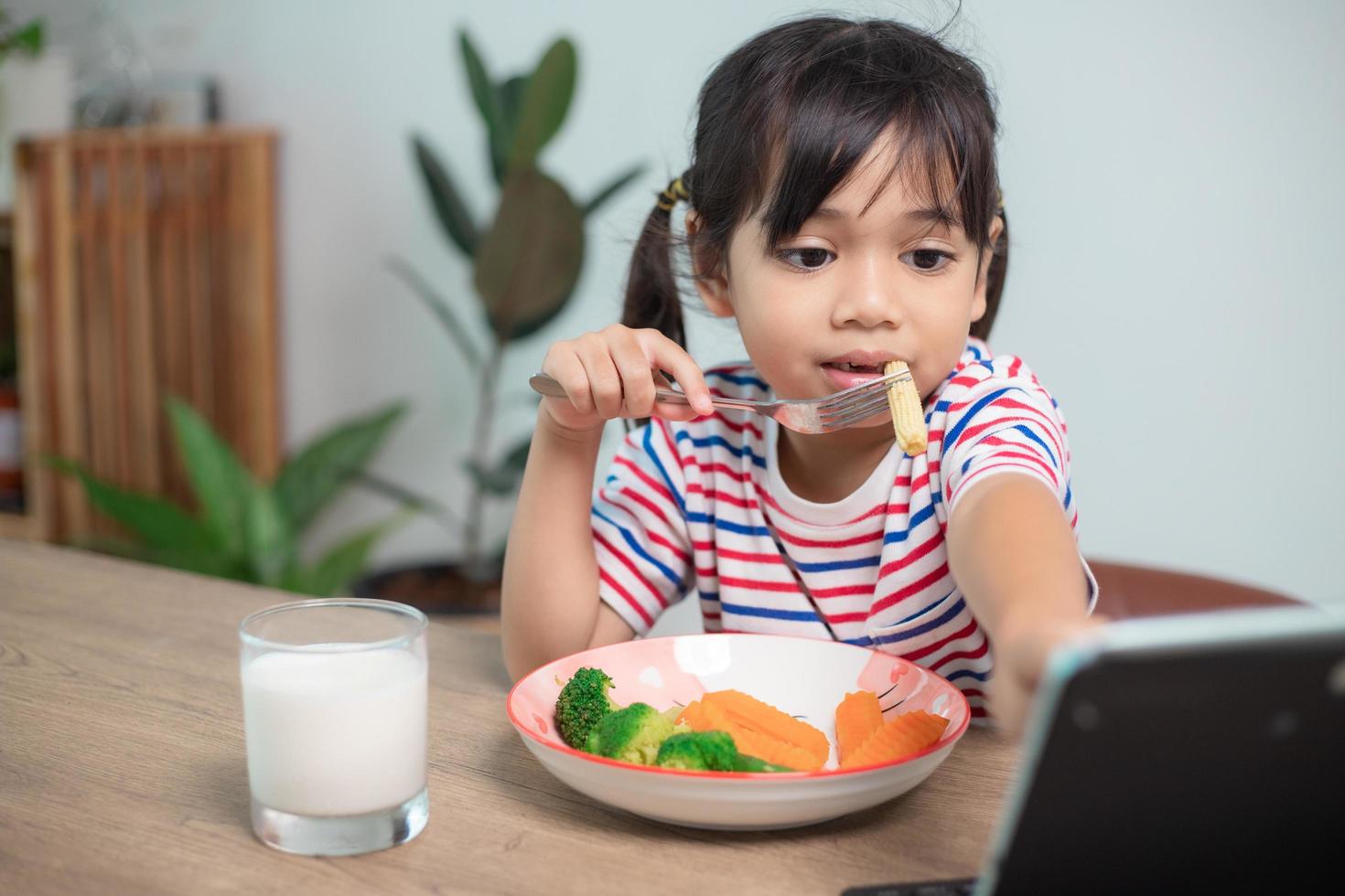 Adorable Asia child girl having lunch while watching a movie from the tablet. A little Asian child eating dinner and eyes are looking cartoon from Tablet. National Eating Disorders Awareness Week. photo