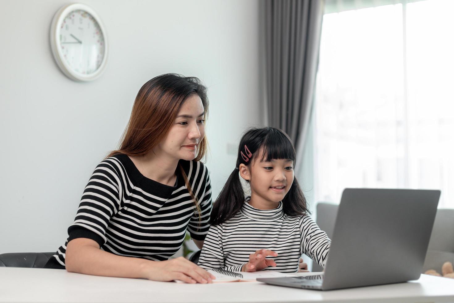 Asian little young girl kid learning online class at home with mother. Preschool child use laptop computer do homework, homeschool from school teacher by digital remote internet with support from mom. photo