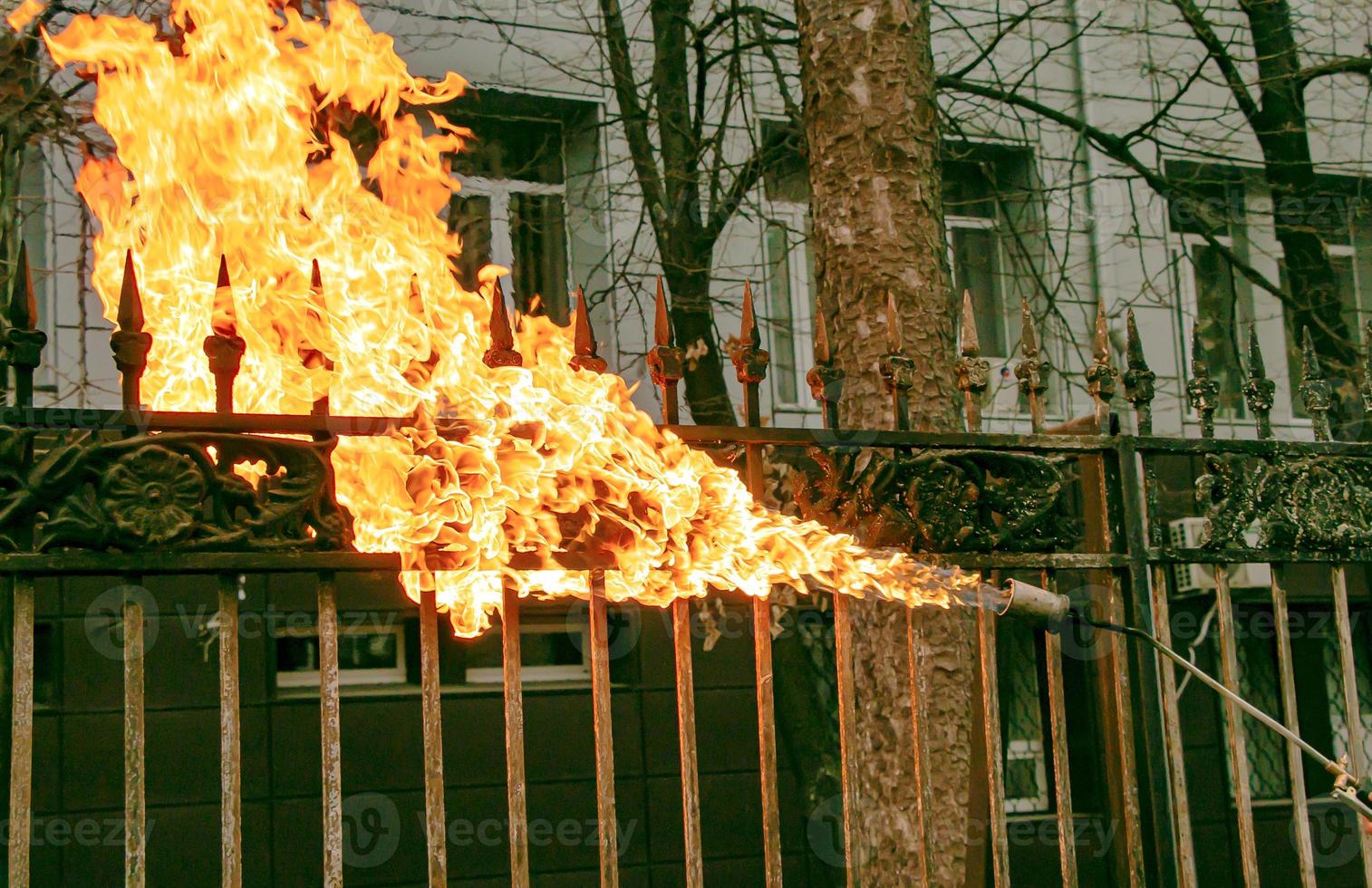 A worker cleans old paint from a metal fence using fire. A man's hand holds a gas burner with a lit flame photo