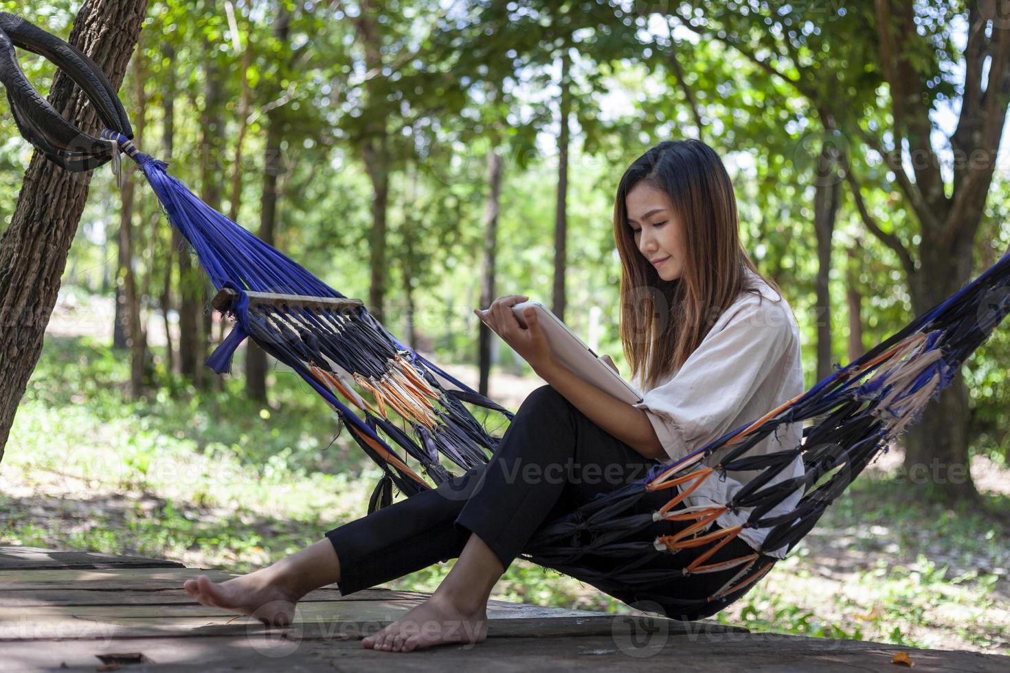 An Asian woman sits in a hammock while working with a tablet under a shady forest. photo