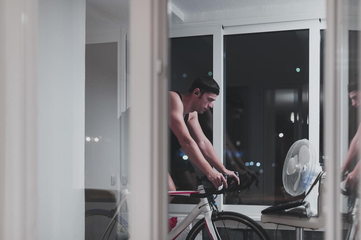 hombre en bicicleta en el entrenador de la máquina que está haciendo ejercicio en el hogar por la noche jugando al juego de carreras de bicicletas en línea foto