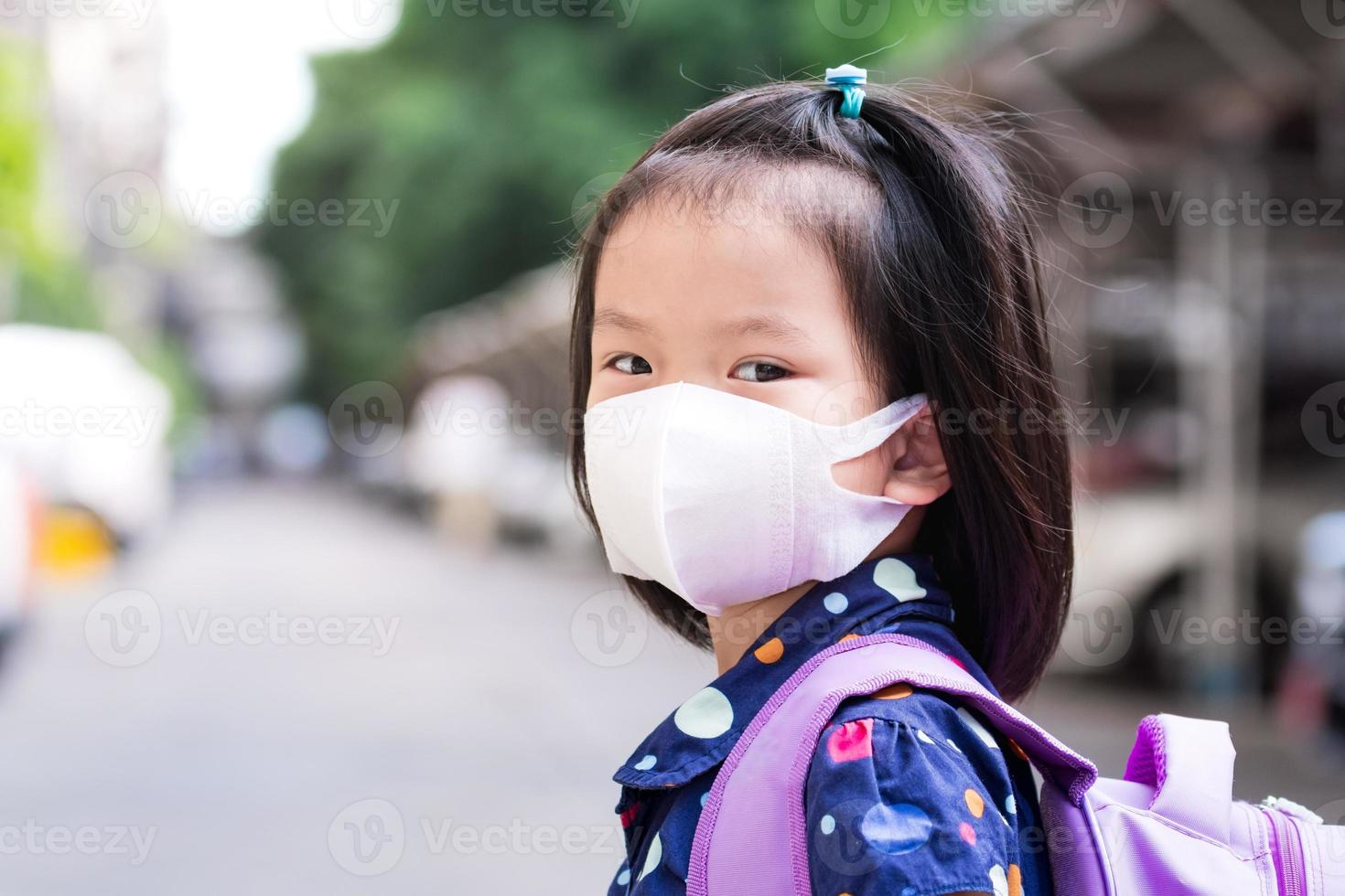 Portrait of child 4-5 years old. Student go to school in summer cause. Little girl wearing white face mask. Sweet smile kid. Children purple backpack. Empty space. photo