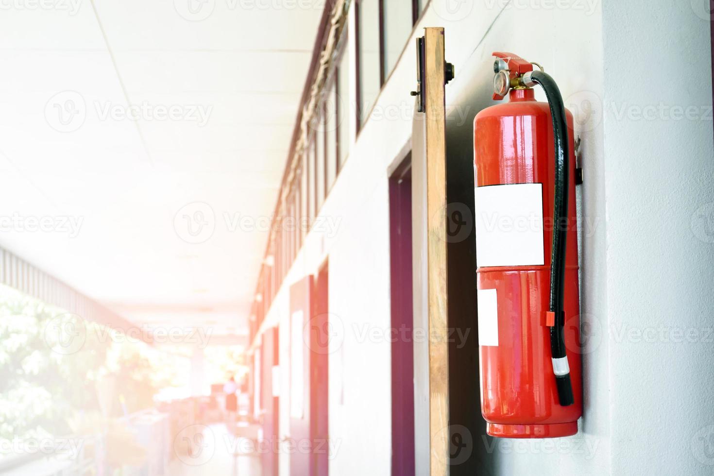 A red fire extinguisher is installed on a white cement wall in the front porch of the building to be used to extinguish a fire in the event of a building fire. photo