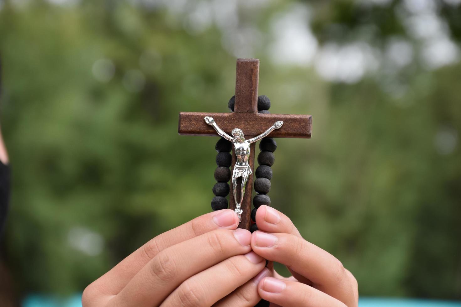 la cruz de madera y el rosario de madera se sostienen en manos de jóvenes católicos asiáticos rezando mientras rezan en el área del parque del templo. foto