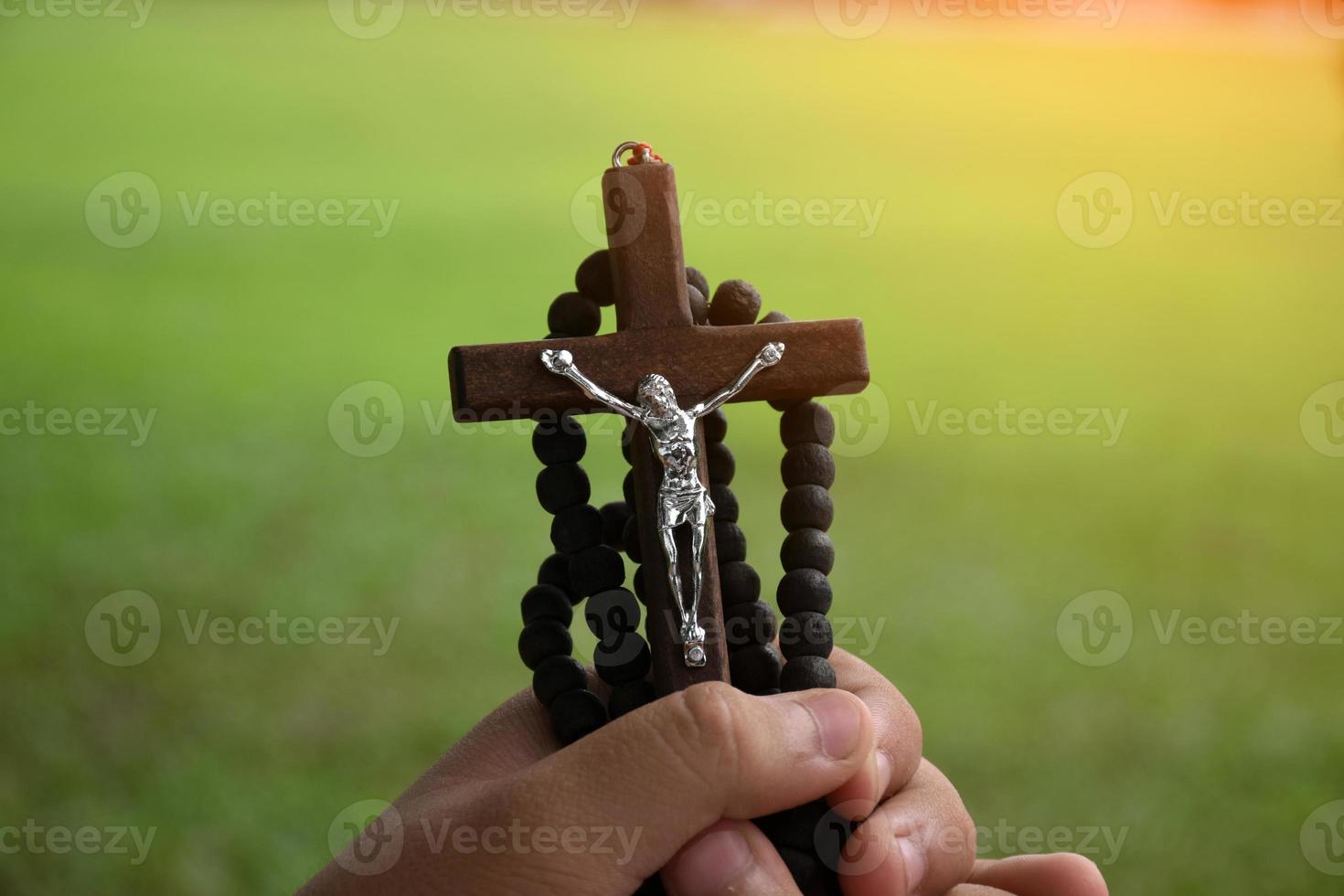 la cruz de madera y el rosario de madera se sostienen en manos de jóvenes católicos asiáticos rezando mientras rezan en el área del parque del templo. foto