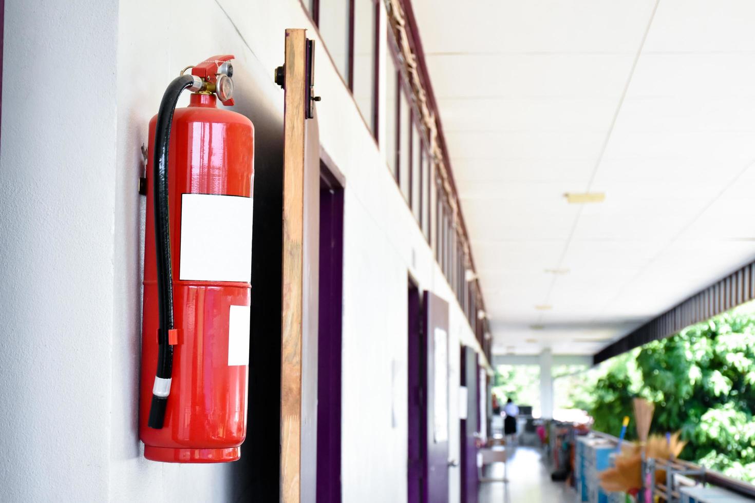 A red fire extinguisher is installed on a white cement wall in the front porch of the building to be used to extinguish a fire in the event of a building fire. photo