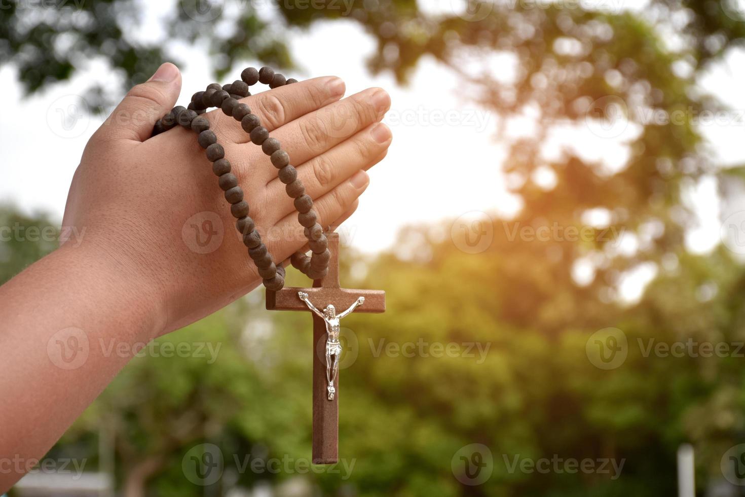 la cruz de madera y el rosario de madera se sostienen en manos de jóvenes católicos asiáticos rezando mientras rezan en el área del parque del templo. foto