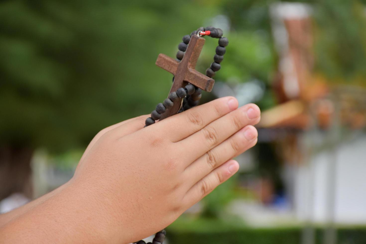 la cruz de madera y el rosario de madera se sostienen en manos de jóvenes católicos asiáticos rezando mientras rezan en el área del parque del templo. foto