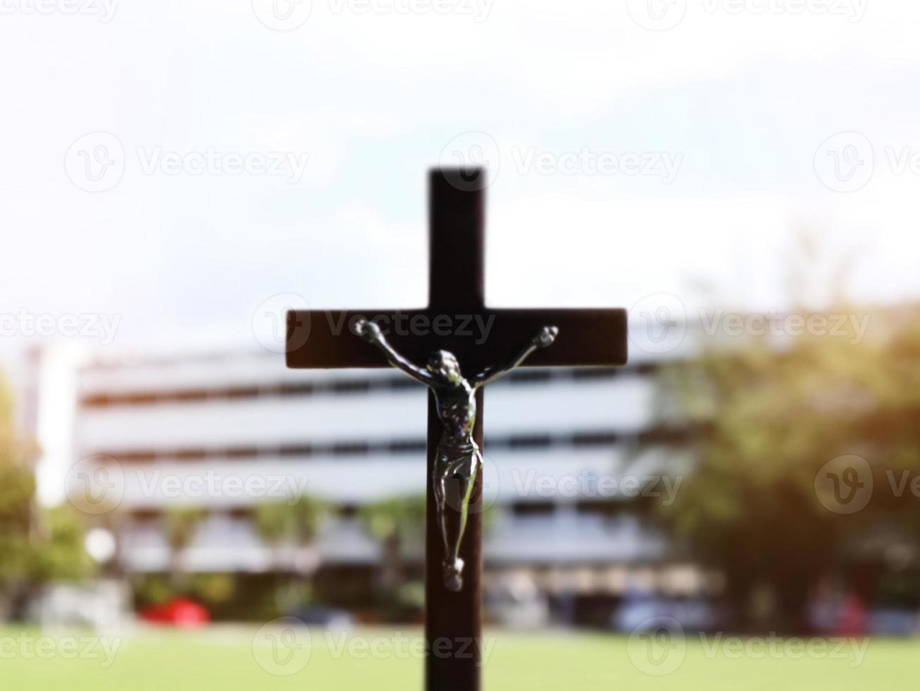 A black wooden cross with a statue of Jesus crucified by his arm. Behind it is the school building of a school in an Asian country, soft and selective focus. photo