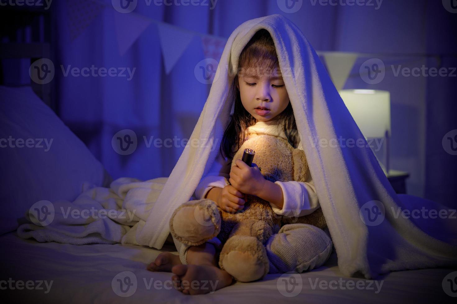 Child asian girl playing teddy bear with flashlight under the blanket on bed in a dark bedroom at night before sleeping, Comfortable children at home concept photo