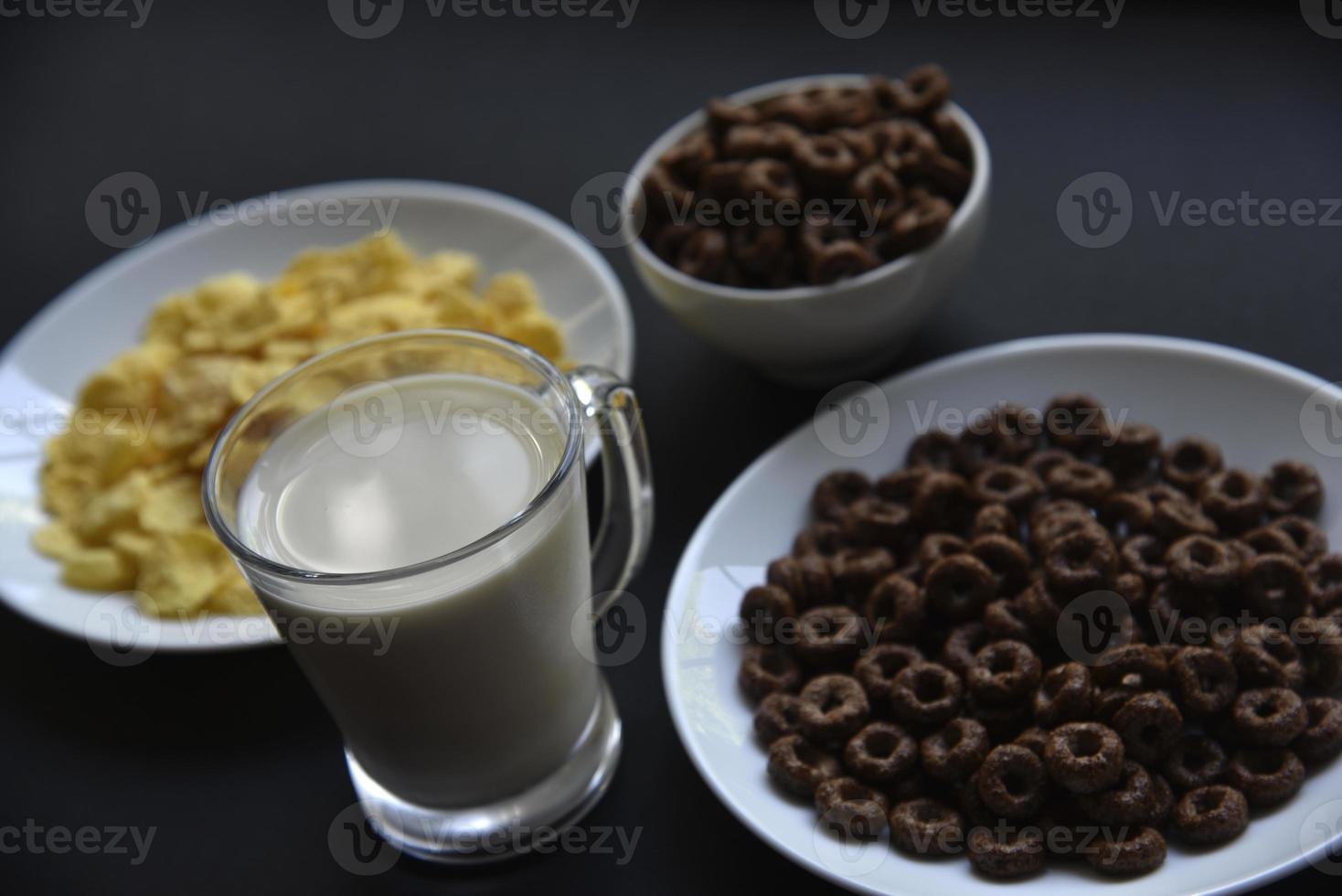 A saucer and a cup of cornflakes and a glass of milk on a black background. Delicious breakfast of cereal with honey and chocolate with milk. photo