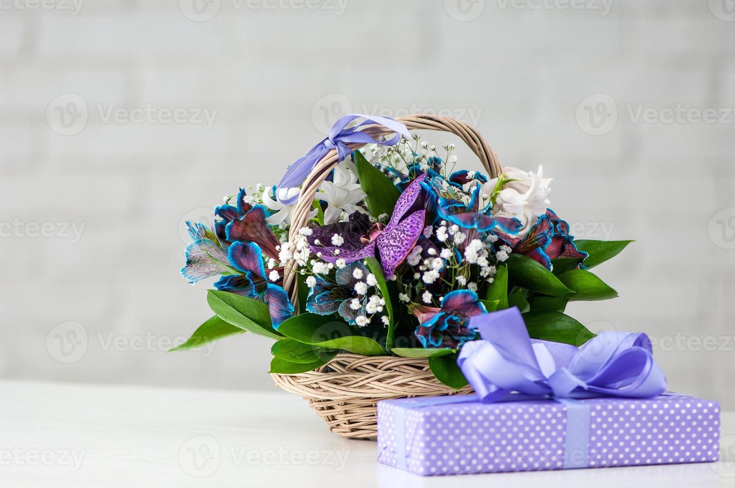 A present next to a composition with orchids, blue alstroemeria, lilies, and in a straw basket photo