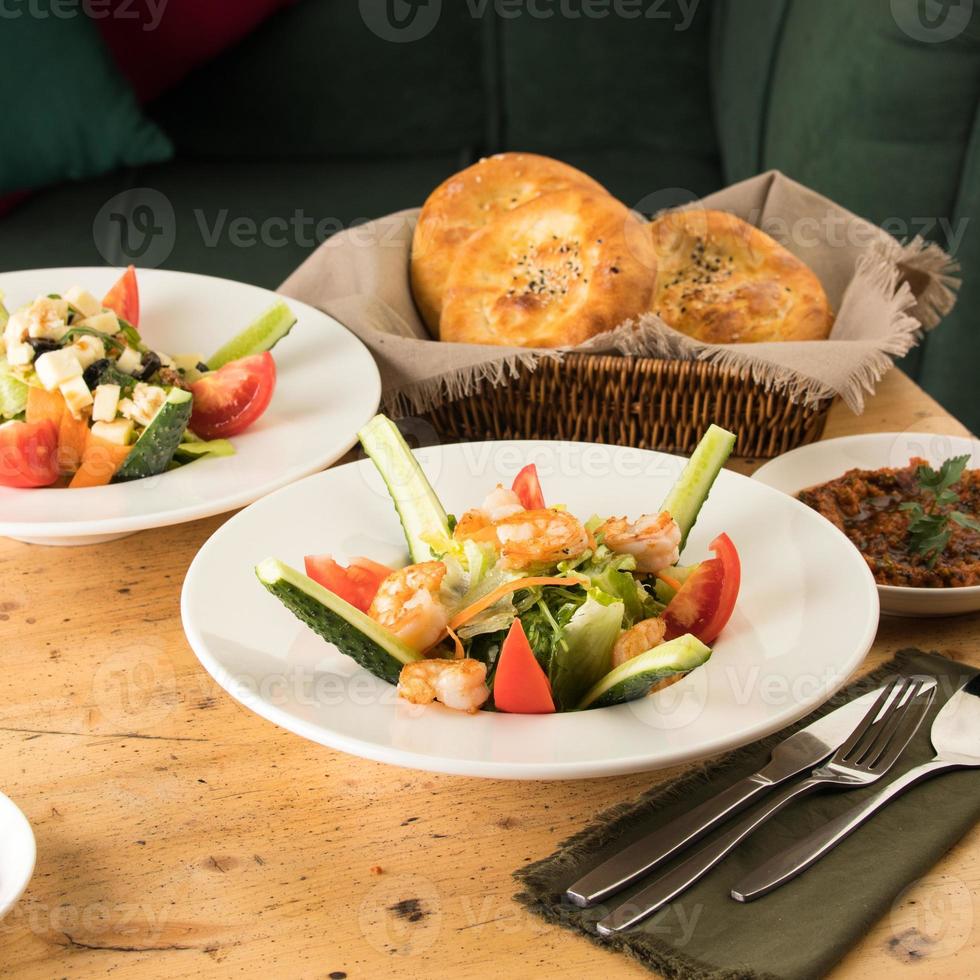 A dining table full of delicious salad on a white plate next to basket of bread photo