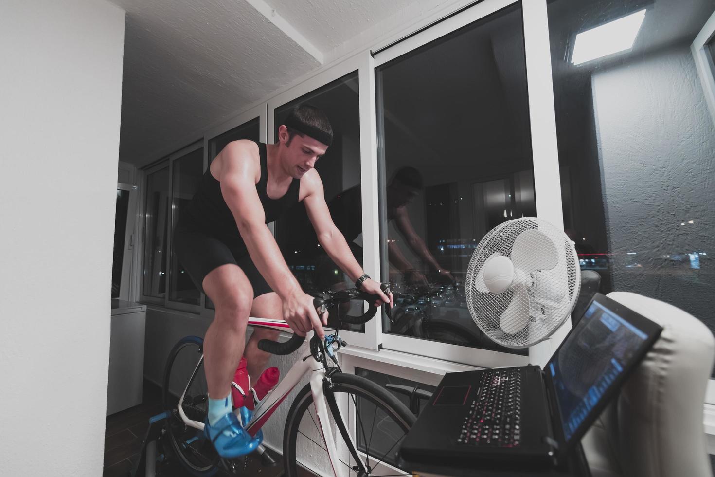 hombre en bicicleta en el entrenador de la máquina que está haciendo ejercicio en el hogar por la noche jugando al juego de carreras de bicicletas en línea foto