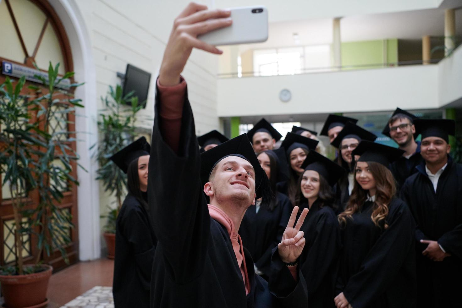 grupo de estudiantes internacionales felices en juntas de mortero y batas de soltero con diplomas tomando selfie por teléfono inteligente foto