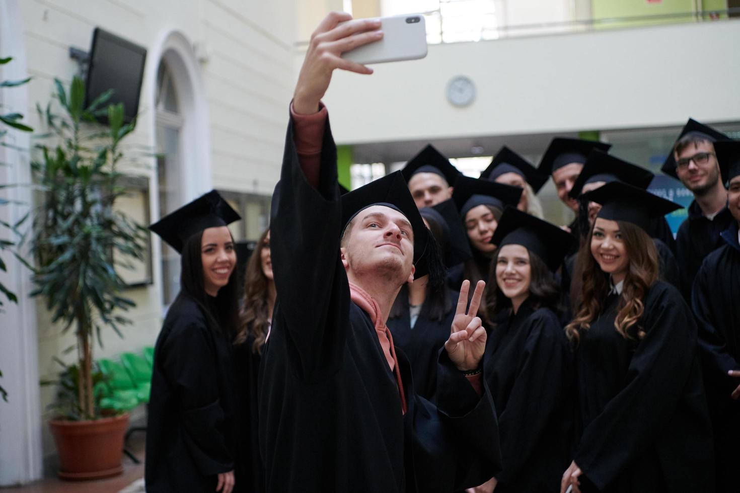 group of happy international students in mortar boards and bachelor gowns with diplomas taking selfie by smartphone photo