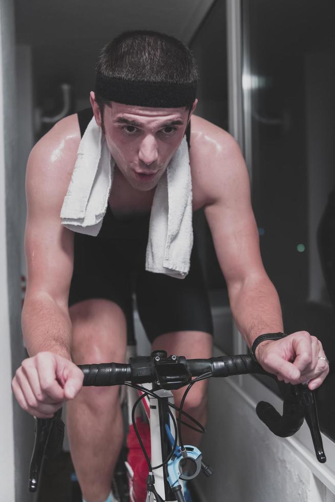 hombre en bicicleta en el entrenador de la máquina que está haciendo ejercicio en el hogar por la noche jugando al juego de carreras de bicicletas en línea foto